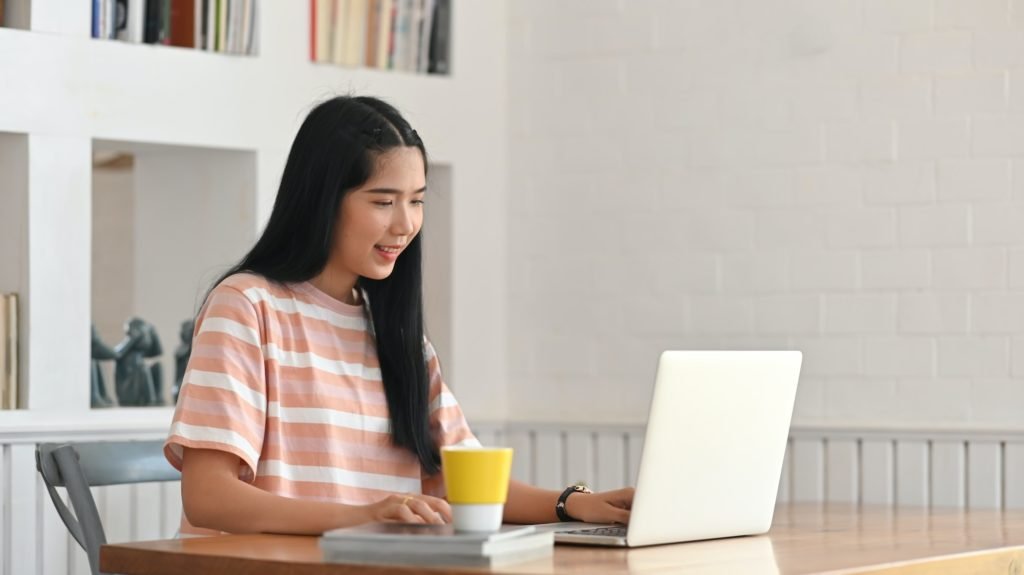 The young woman is typing on a computer laptop while sitting at the wooden working desk.